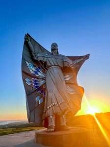 The Dignity Statue in Chamberlain, South Dakota, standing tall at sunrise, with golden light highlighting the Native American woman’s flowing star quilt and intricate details.